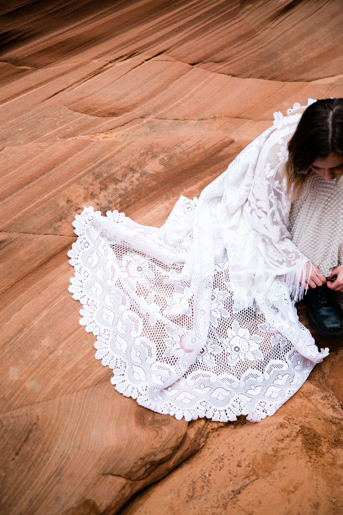 During her elopement in Arizona a bride ties her boots. Her lace dress fans out beside her, stark white against the red rock of the canyon.