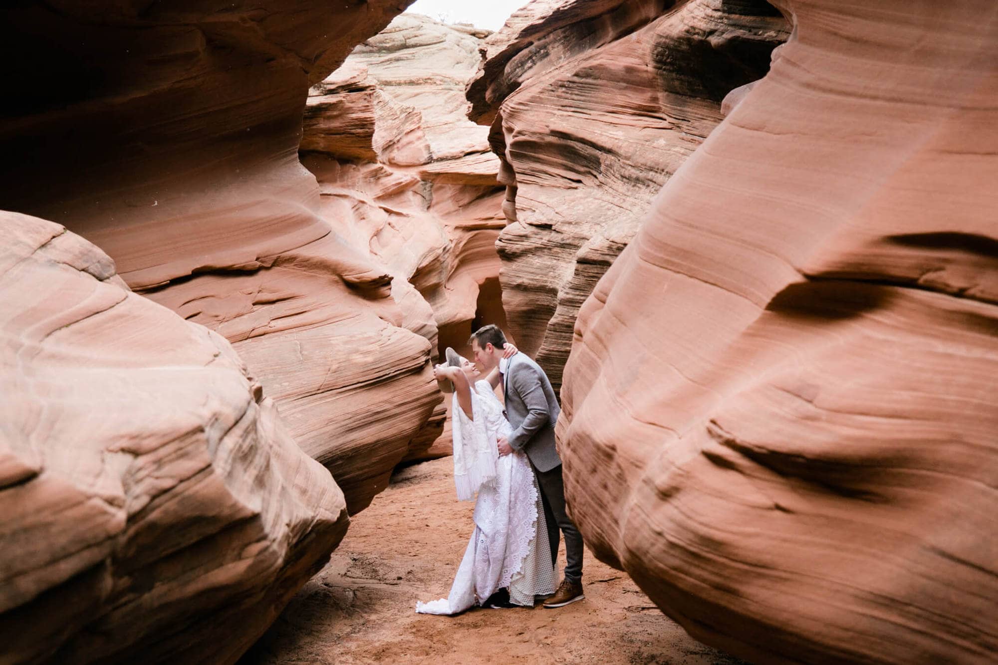 During an elopement in Arizona, the groom dips the bride, surrounded by the slot canyon walls. Laughing, the bride grabs her hat.