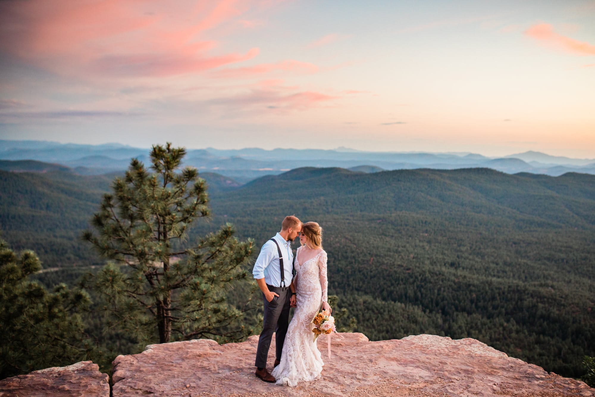 Having just eloped in Arizona, a bride and groom stand near a cliff's edge. The sunset sky turns pink behind them.