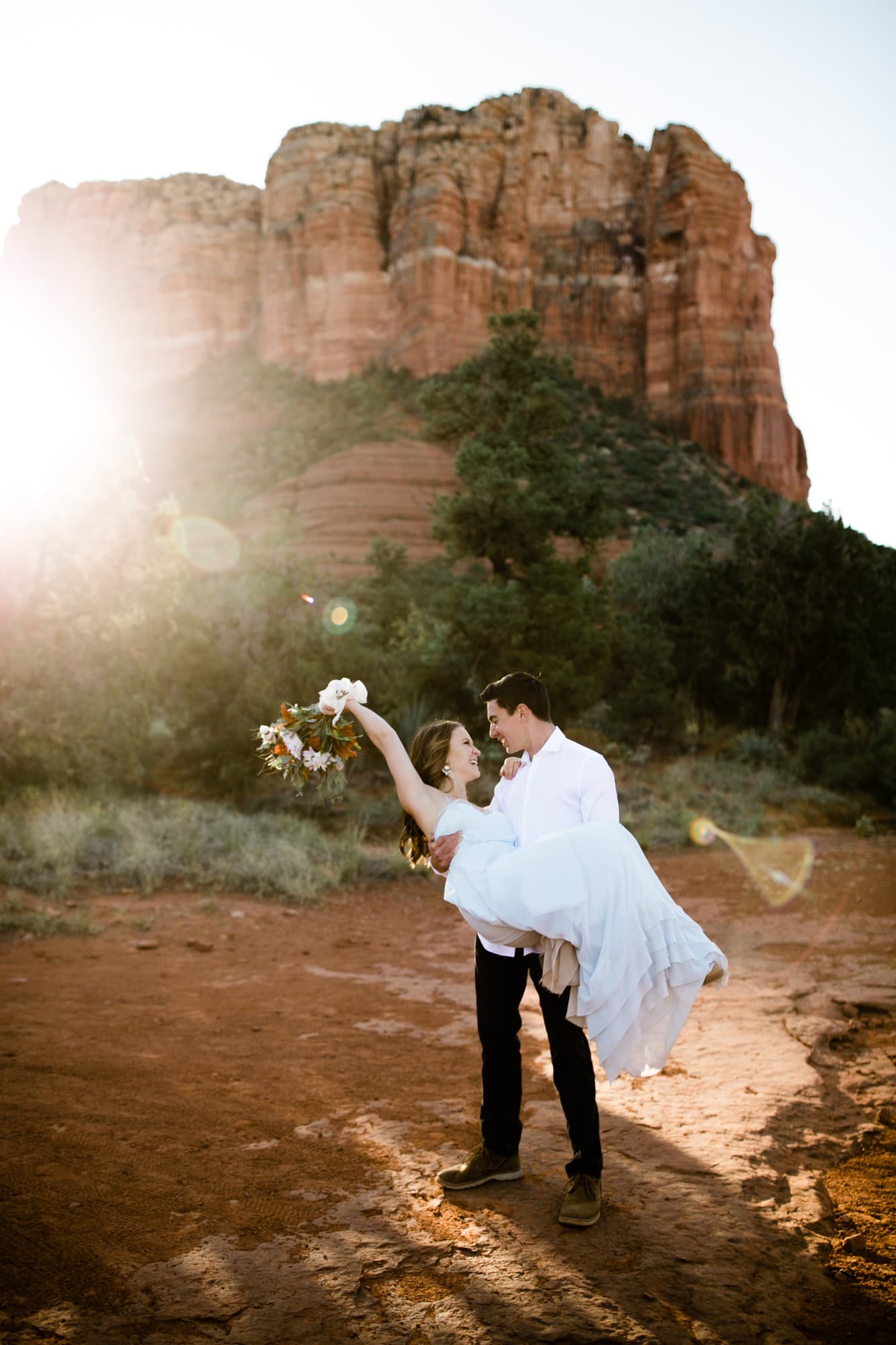 The groom picks up the bride. She holds her bouquet over her head and the sun shines behind them.