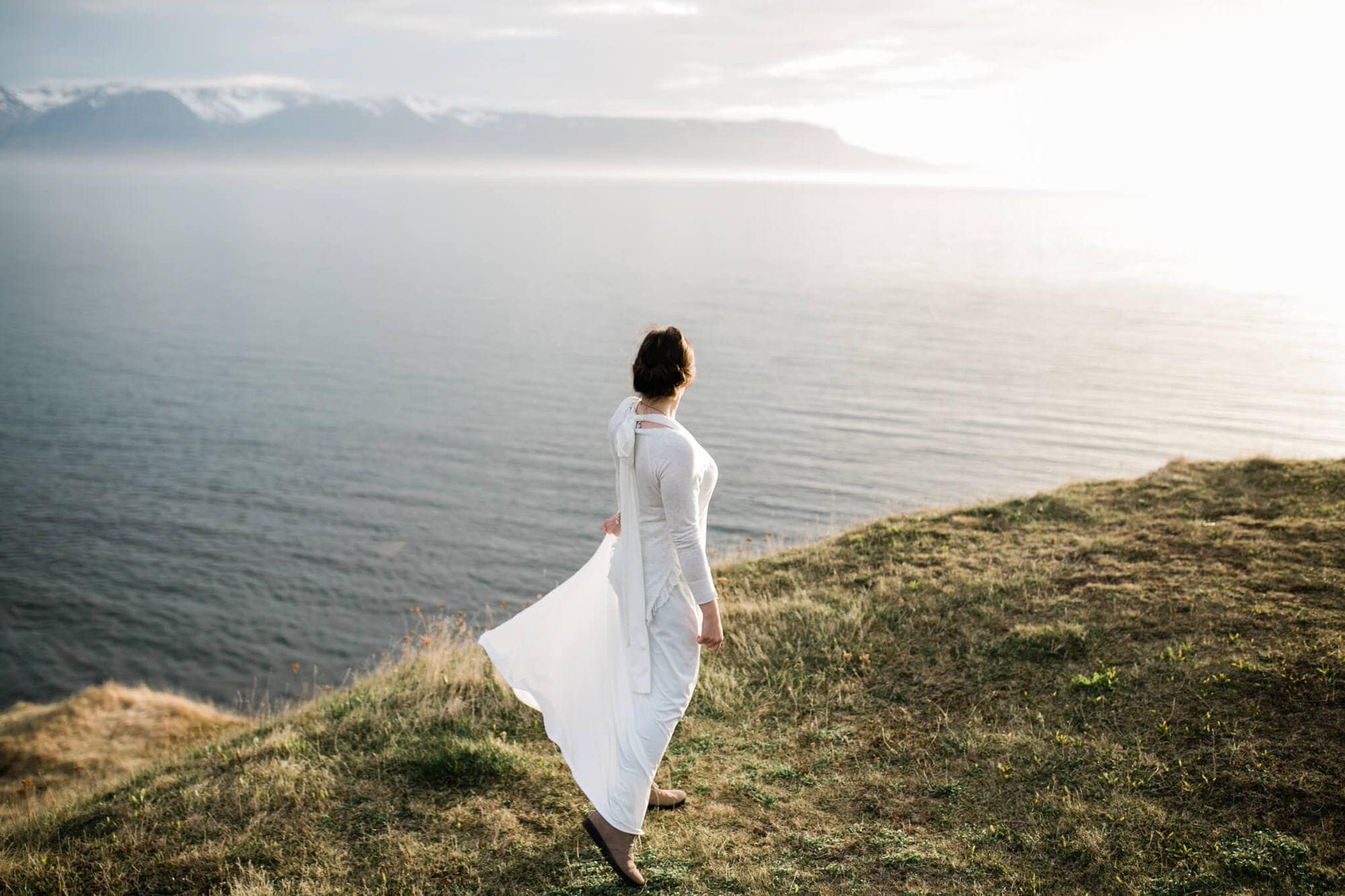A bride looks out over a sea cliff at sunset.