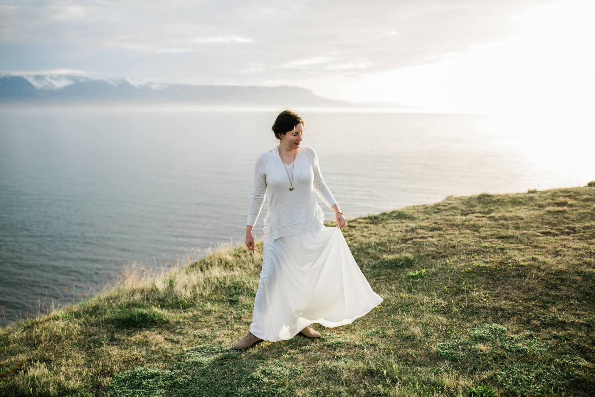 A bride stands on a cliff overlooking the ocean at sunset in Iceland.