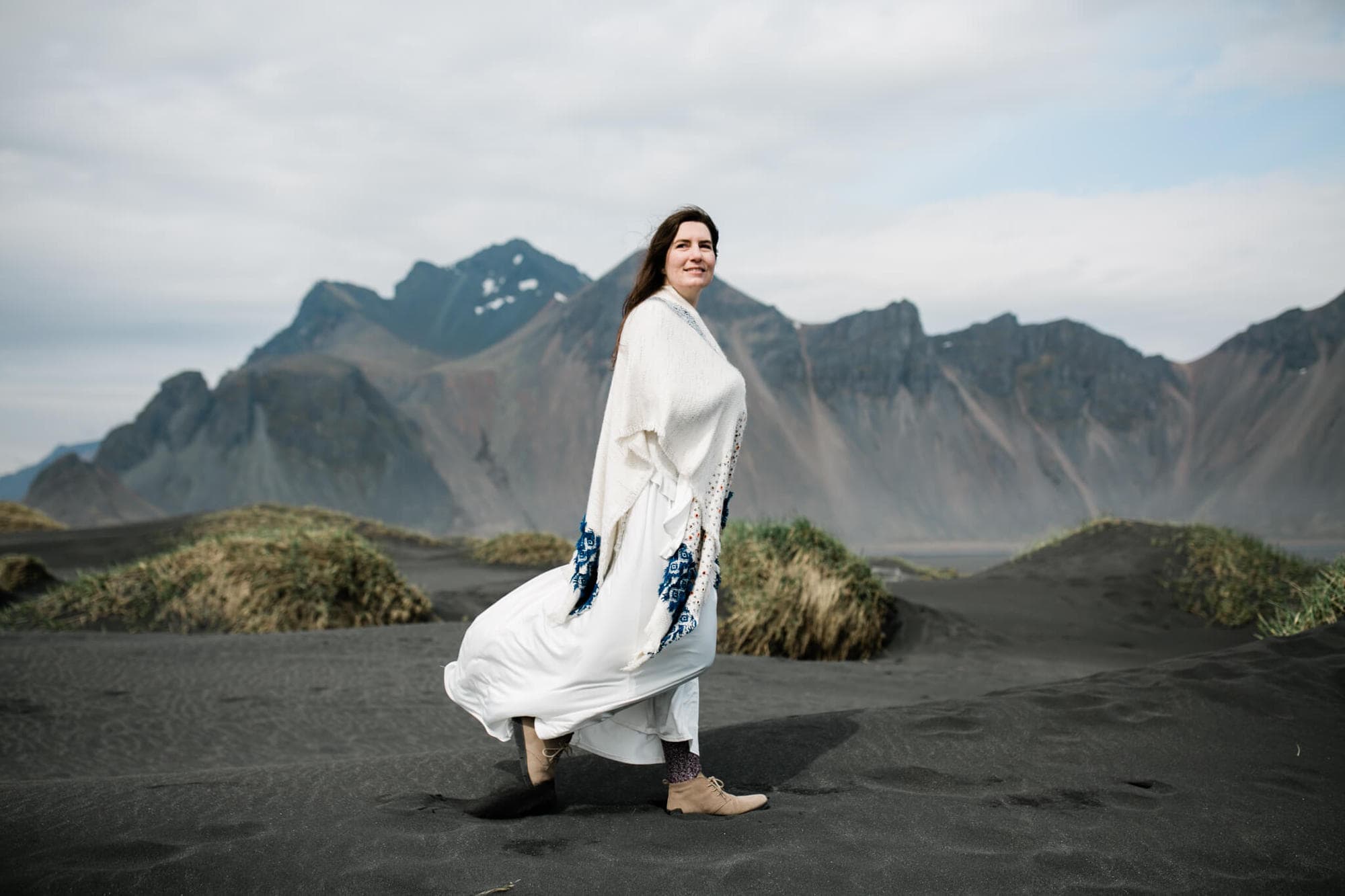 A bride walks along a black sand beach in Iceland. Wind blows her hair and dress.