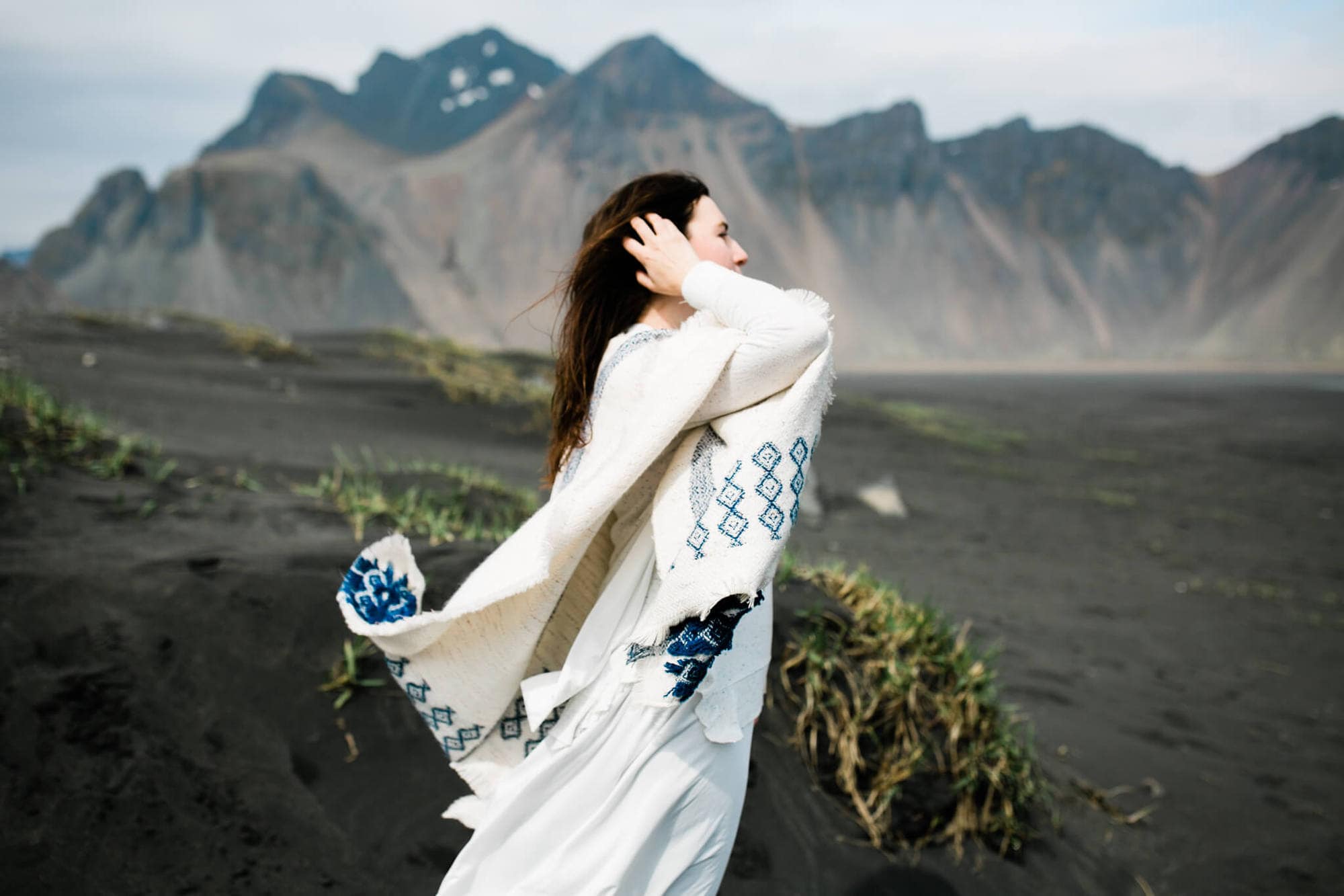 Standing on a black sand beach, a bride fixes her hair as the wind catches her sweater cape.