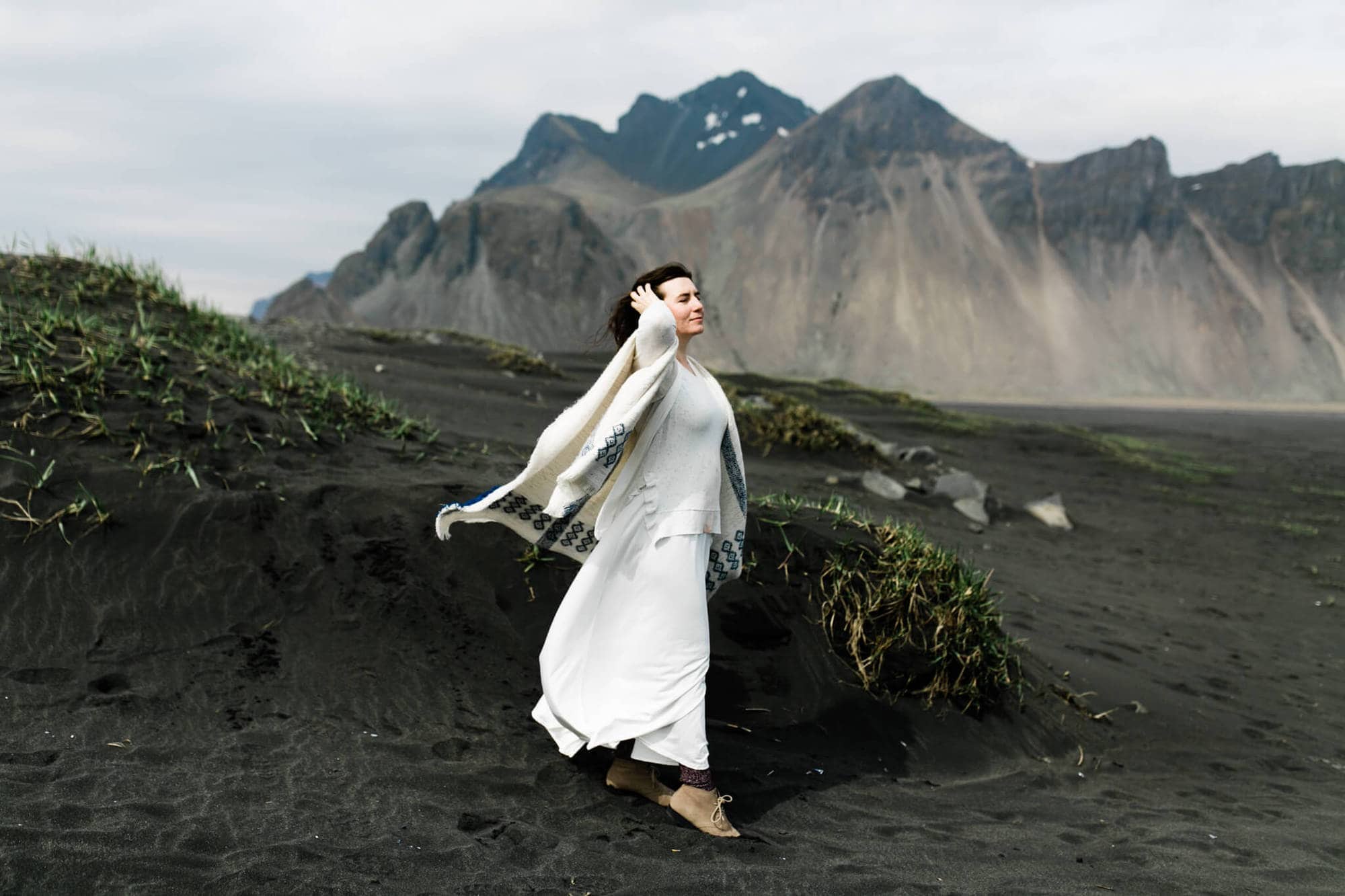 A bride stands among black sand dunes in Iceland. Wind catches her dress and sweater cape.