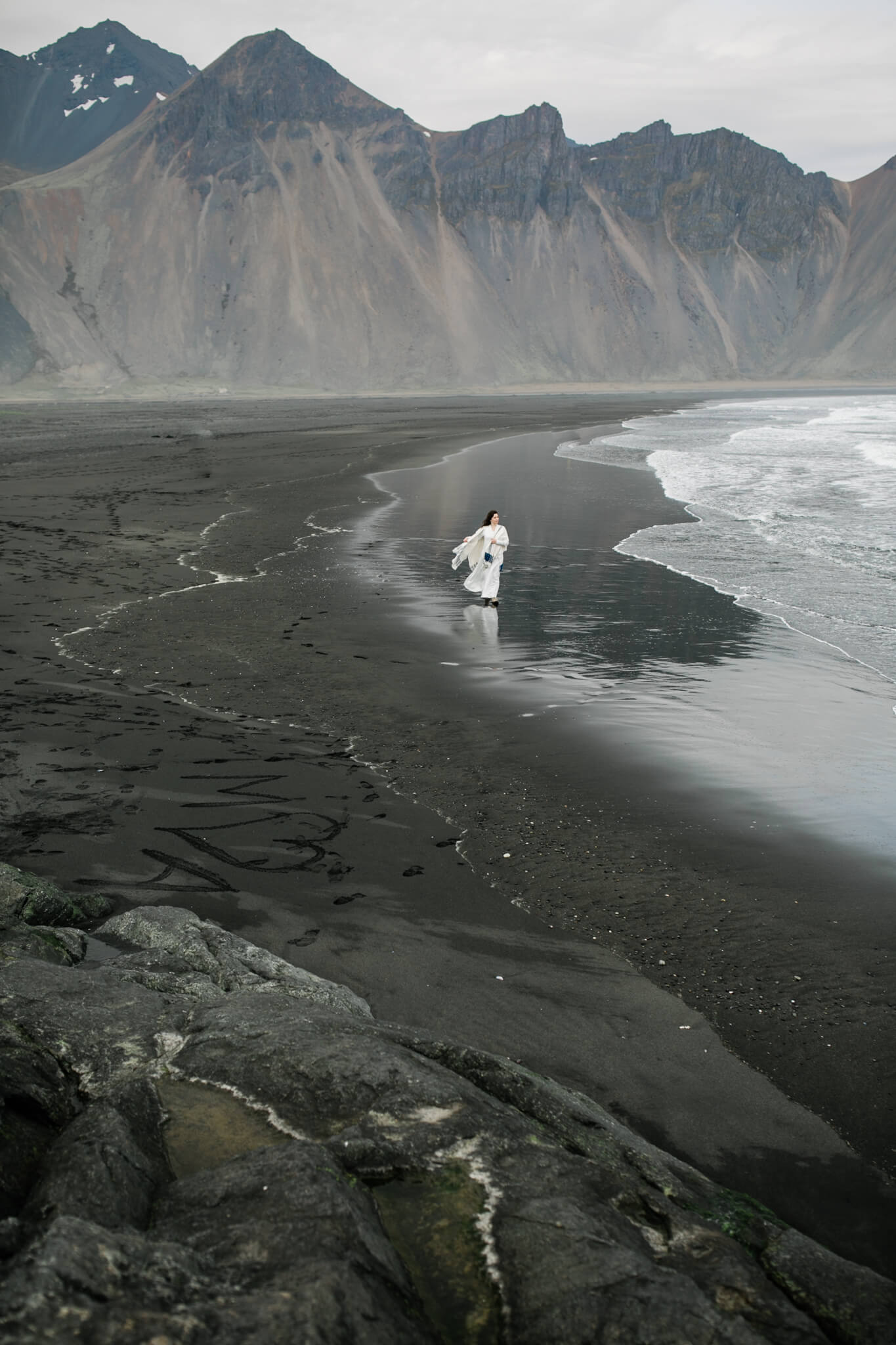 A tiny bride, dwarfed by huge mountains behind her, walks along a black sand beach.