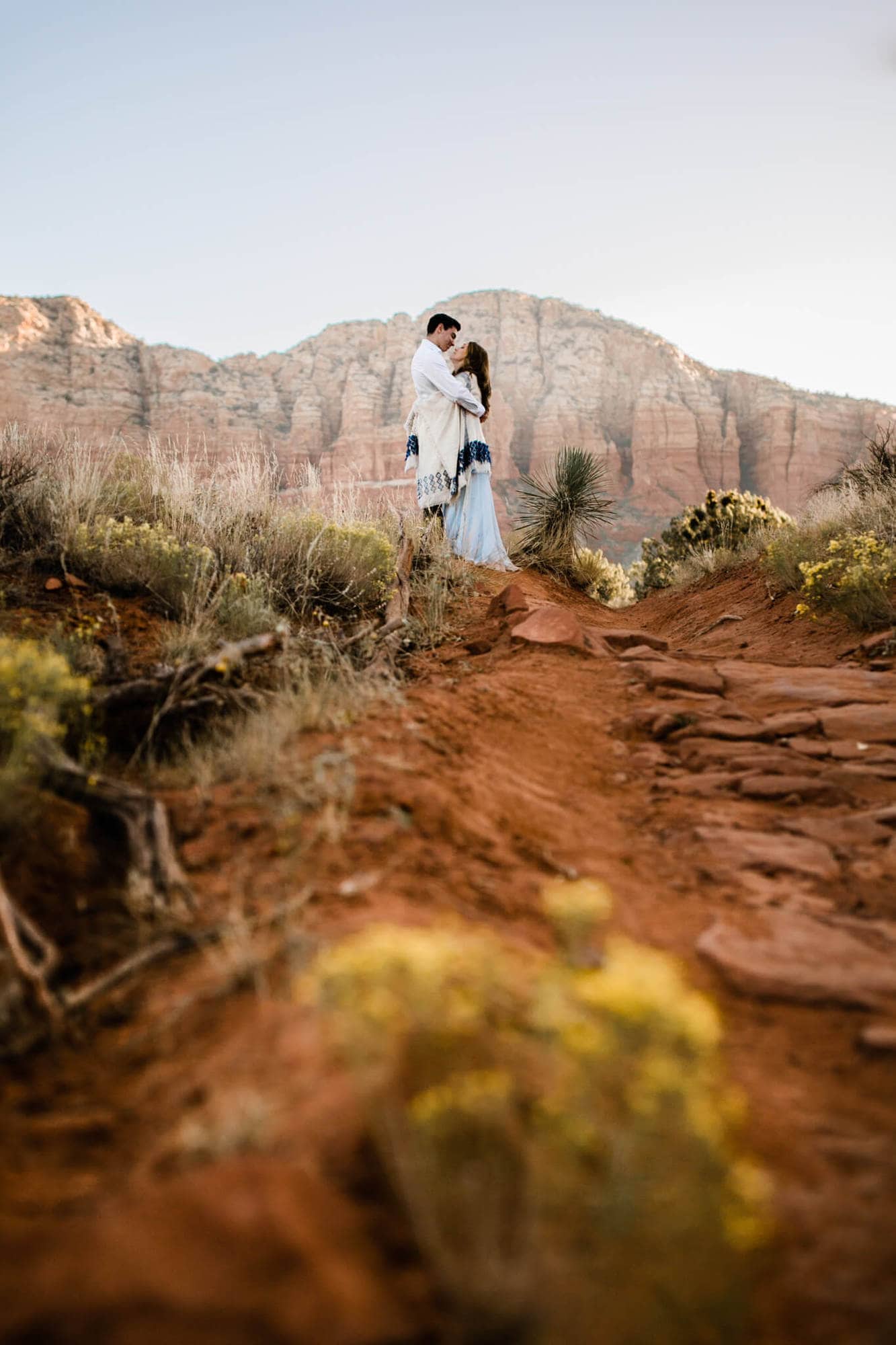 Standing on a hill and framed by the red mountains behind them, the bride and groom hug.