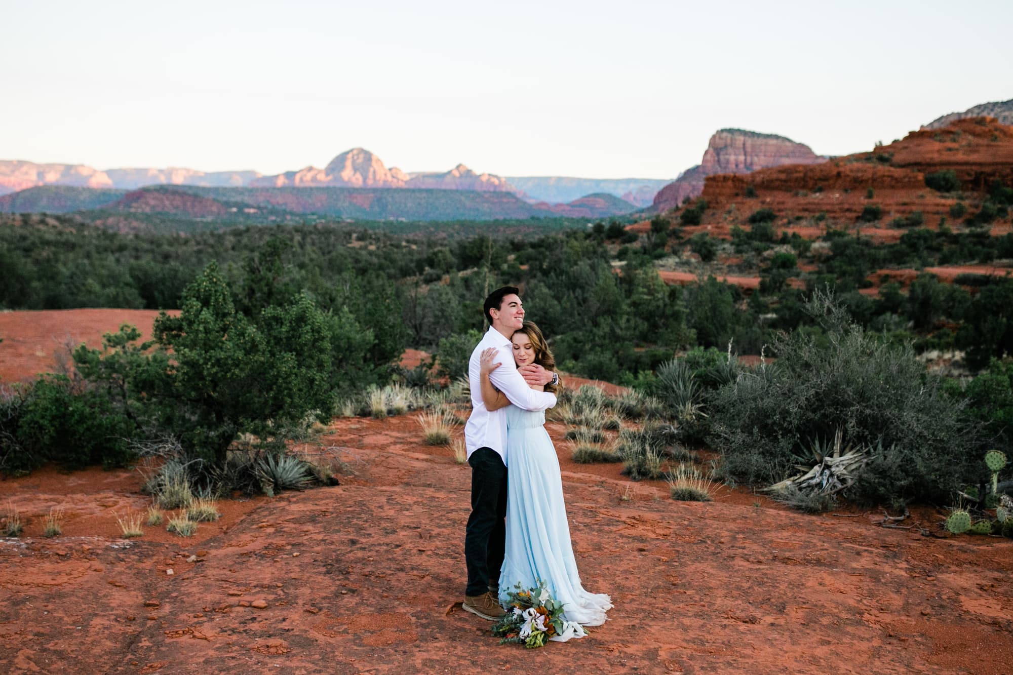 Newly married, the bride and groom hug with the red rock mountains in the distance.