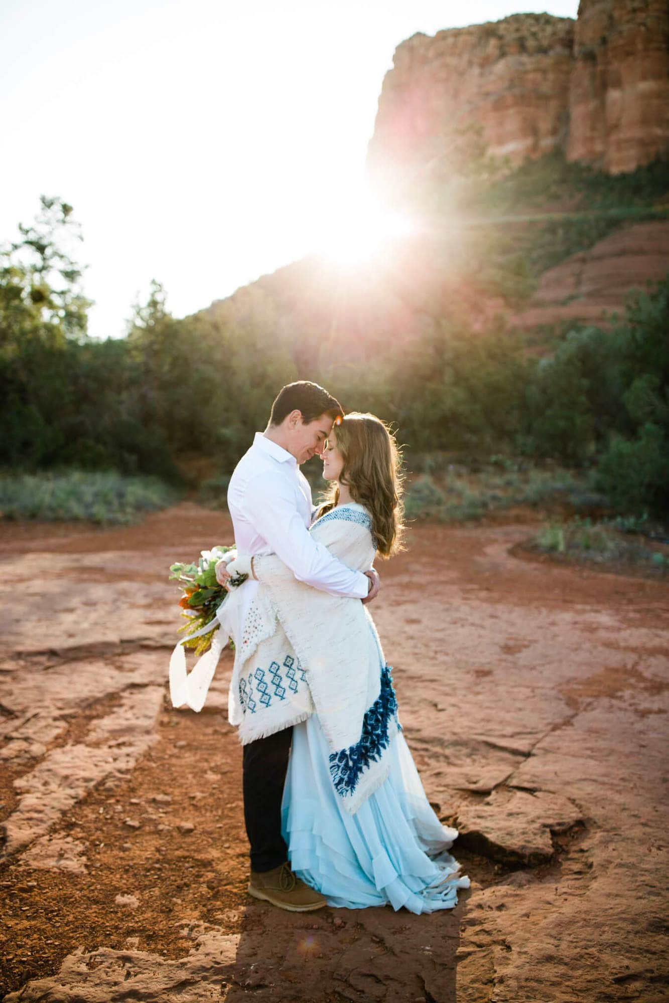 Couple hugs, touching foreheasds, while the sun bursts from behind Courthouse Butte behind them.