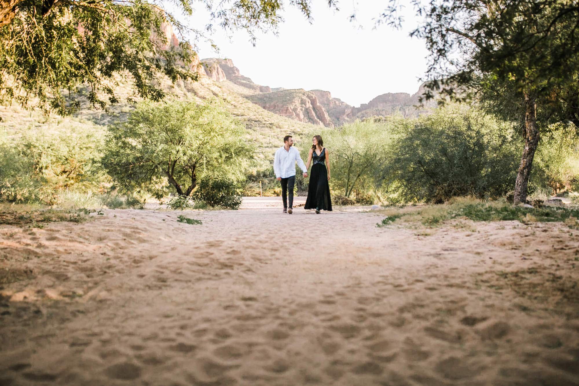 The couple walks on a sandy pathway through some trees. The  desert mountains glow behind them.