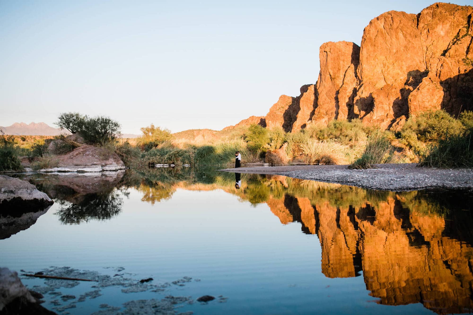 The adventurous couple looks tiny next to the tall, sunlit desert cliffs. Their reflection is perfect on the still water.