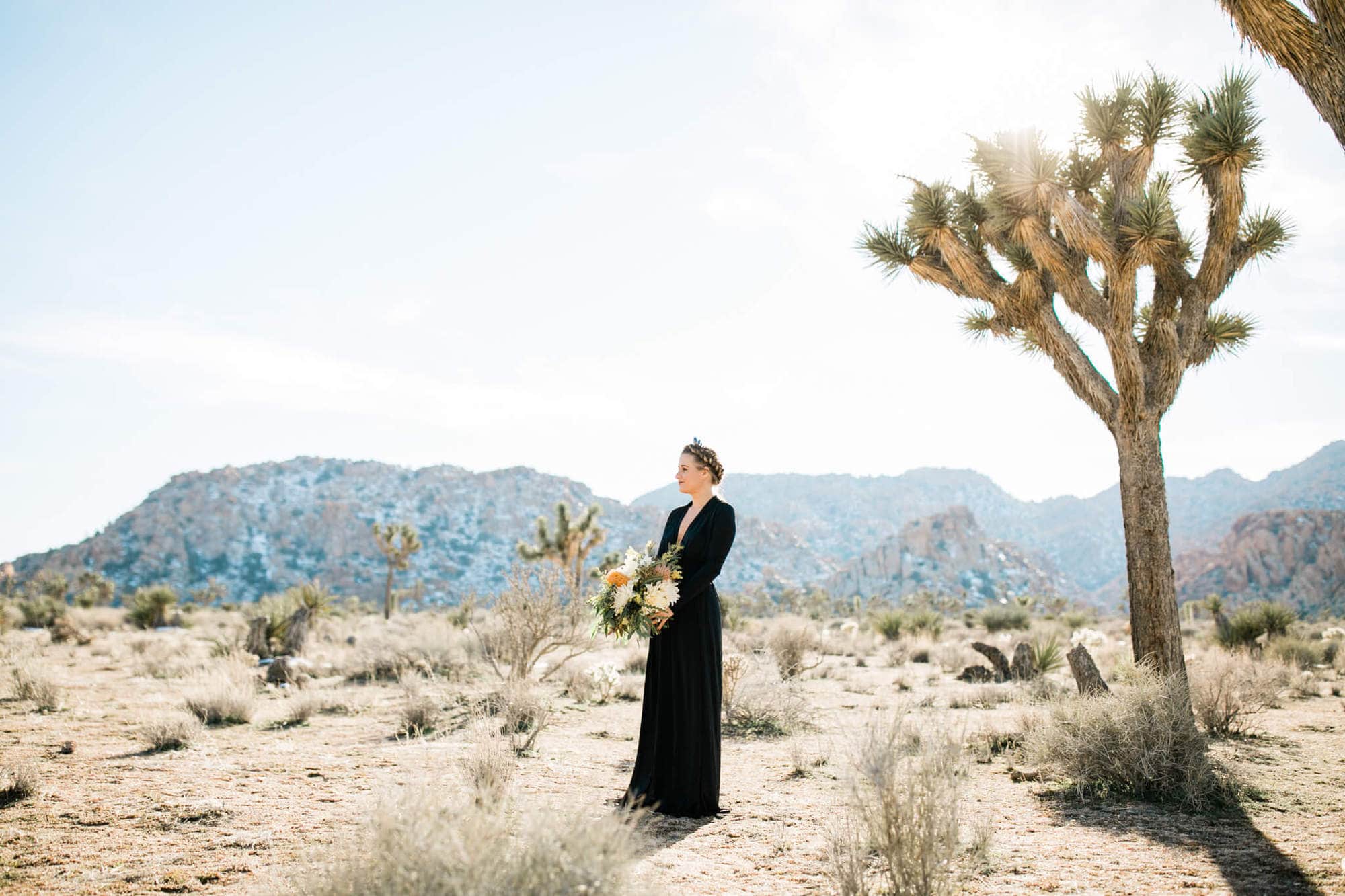 A bride in a black wedding dress holds a beautiful bouquet, standing near a Joshua Tree with mountains in the distance.