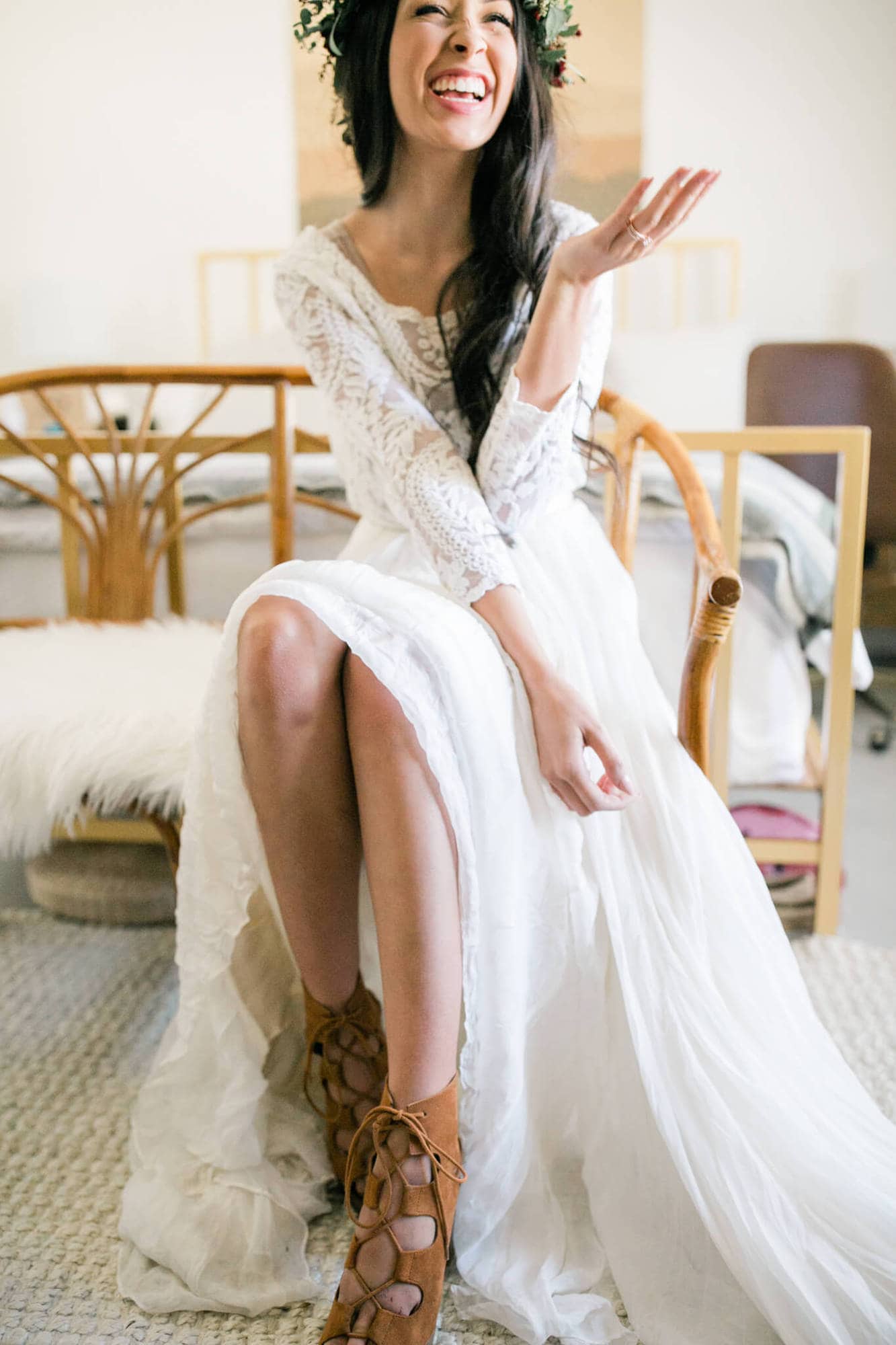 In her wedding dress, the bride sits on a bench in front of her hotel bed and laughs.