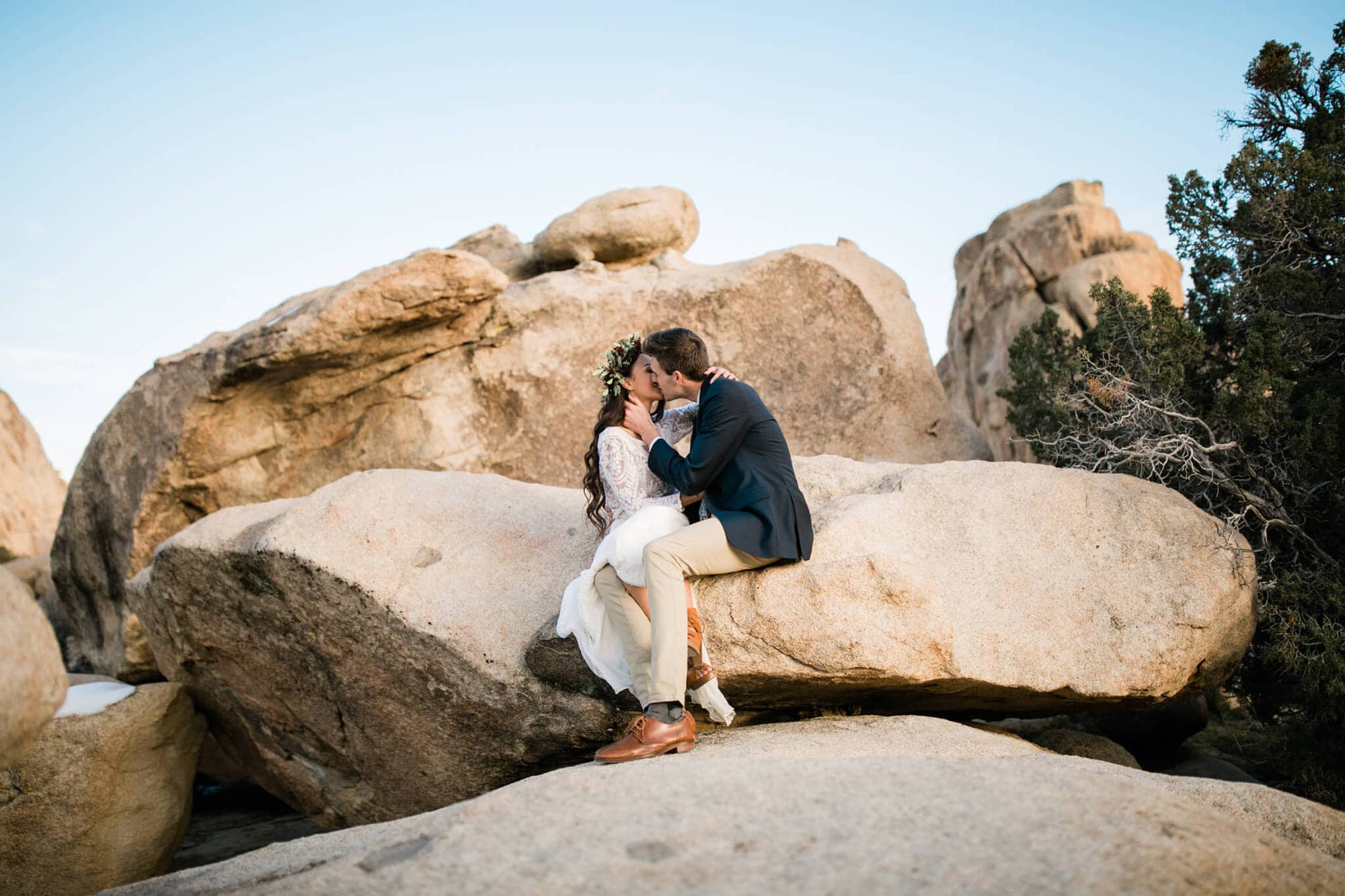 The bride and groom sit on Joshua Tree's boulders and kiss. 