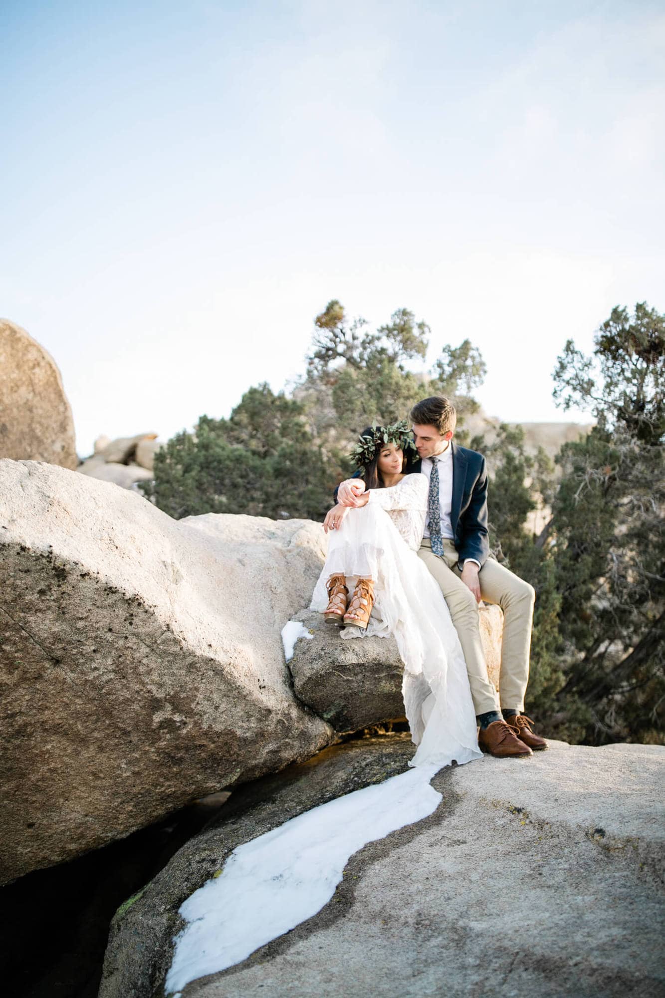 The bride and groom sit atop Joshua Tree's desert boulders. The brides wedding dress blends into a pile of snow, looking like a long train.