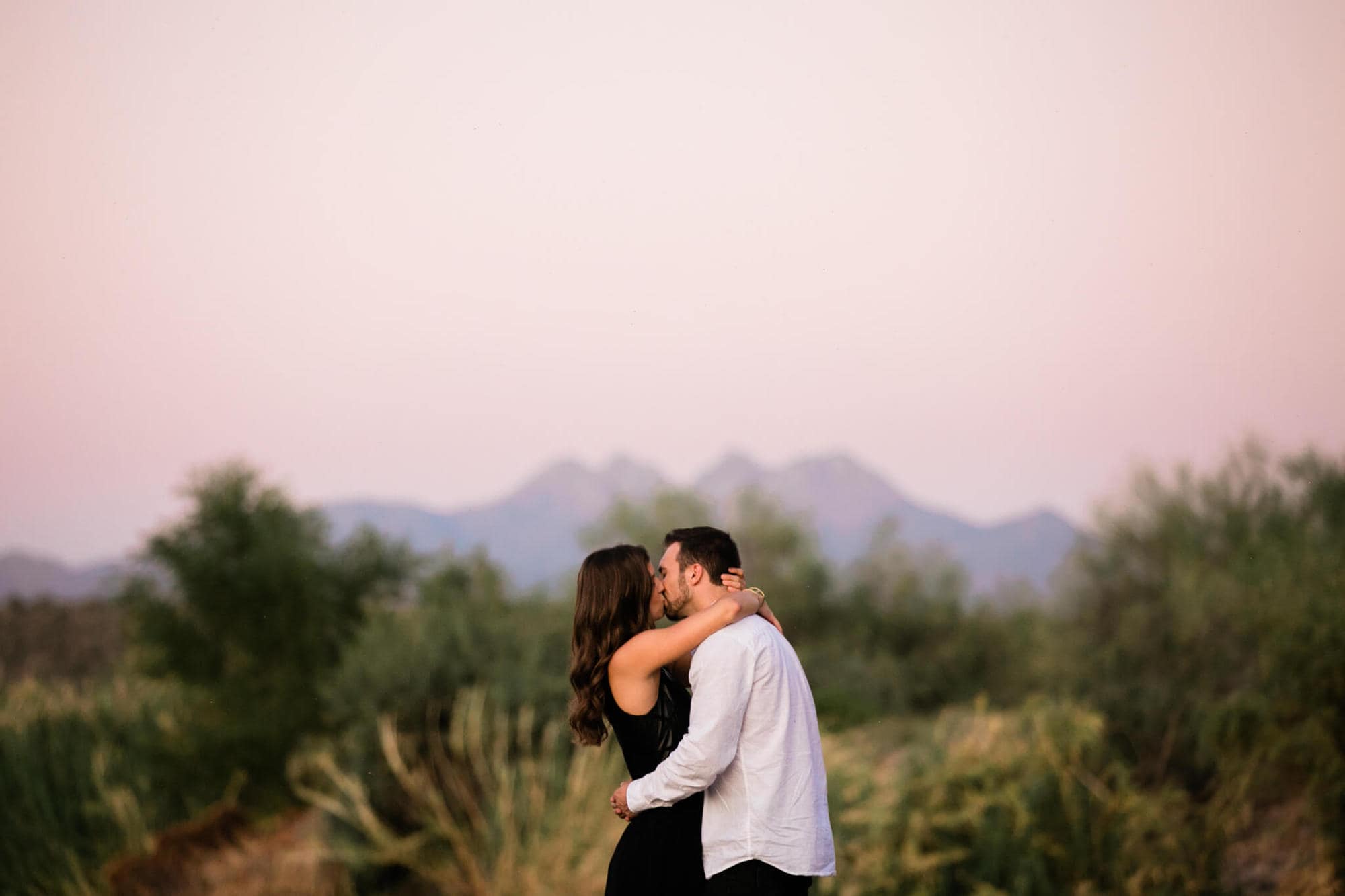 Nicole and Logan kiss with the pink dusk sky behind them and Four Peaks in perfect view.