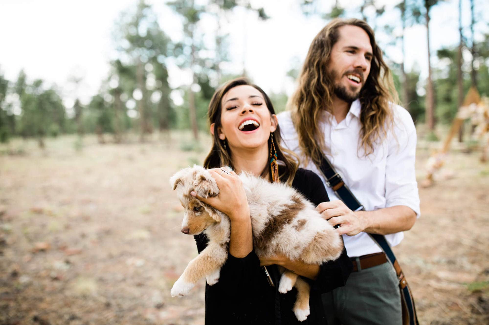 The couple holds a puppy and laughs during the forest engagement photos.