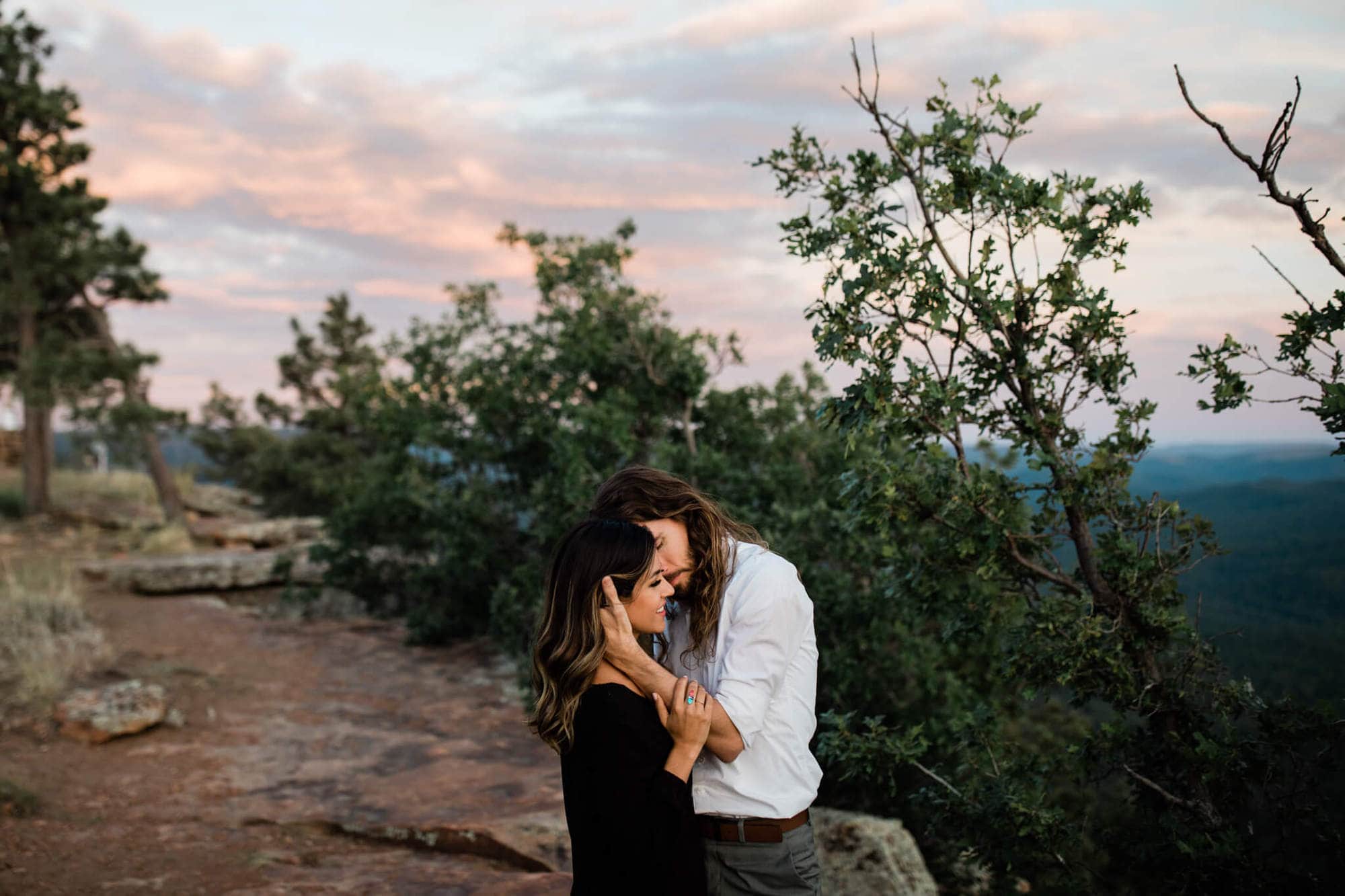 In the fading sunset light, the couple stands at the edge of the cliff during their forest engagement photo session.
