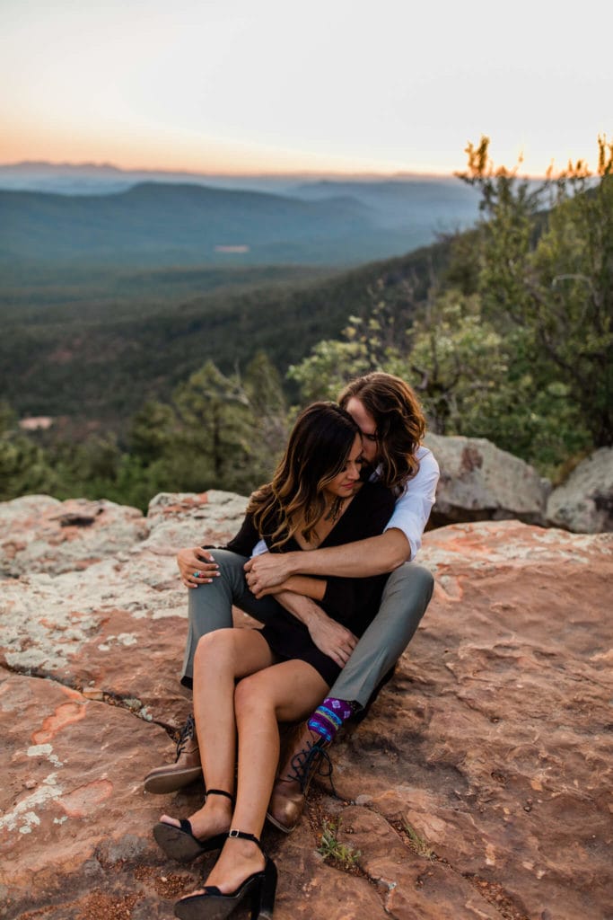 Couple sit on the edge of the Mogollon rim during the adventurous engagement photos. The sun sets behind them.