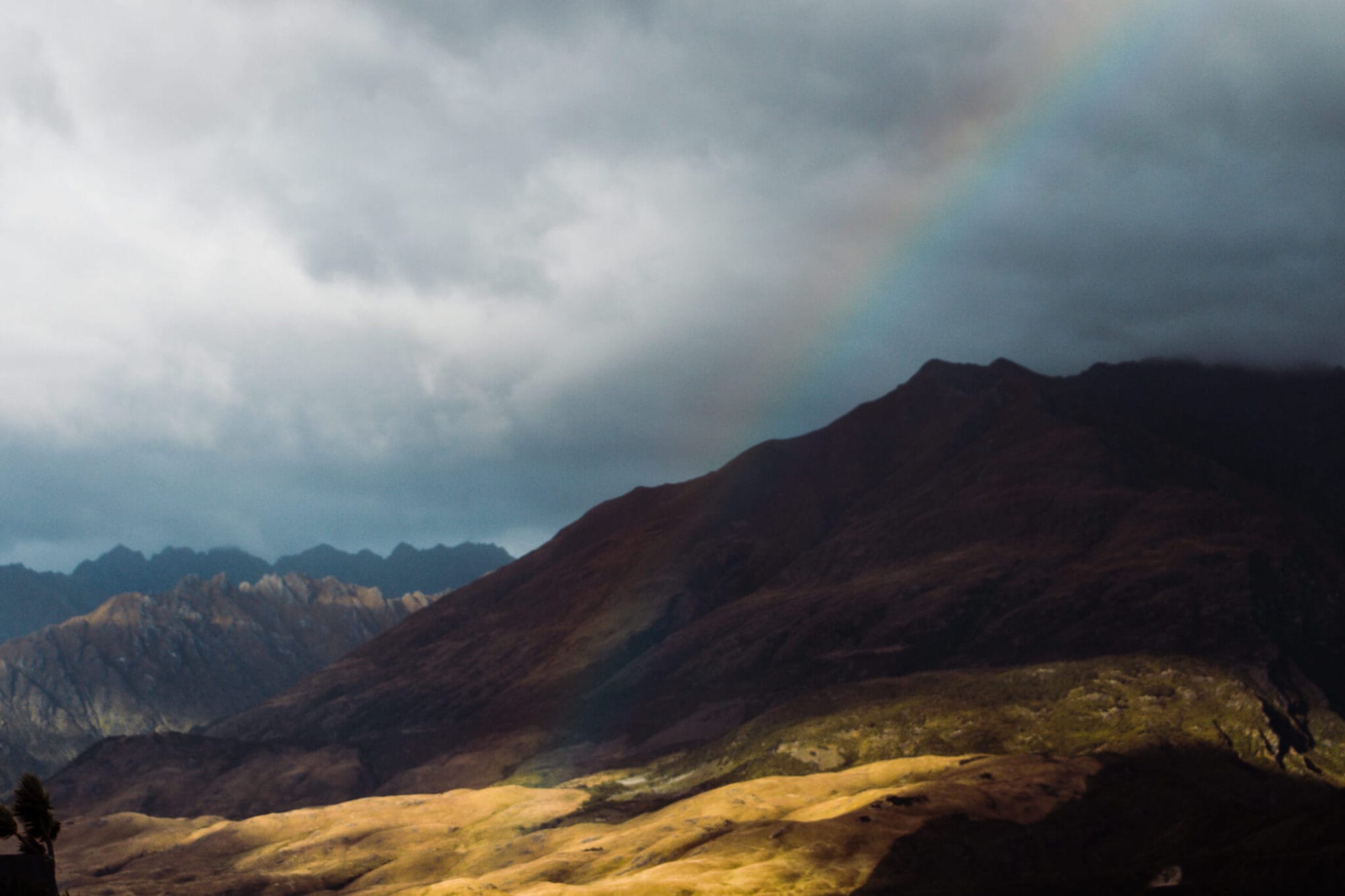 A rainbow appears over the mountains of New Zealand's South island. The sun exposes patches of yellow grass on the hillside.