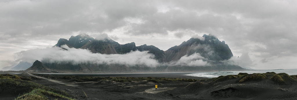 A girl in a yellow jacket stands, tiny in the massive sprawl of black sand dunes. On the horizon, clouds cloak over the dark mountains.