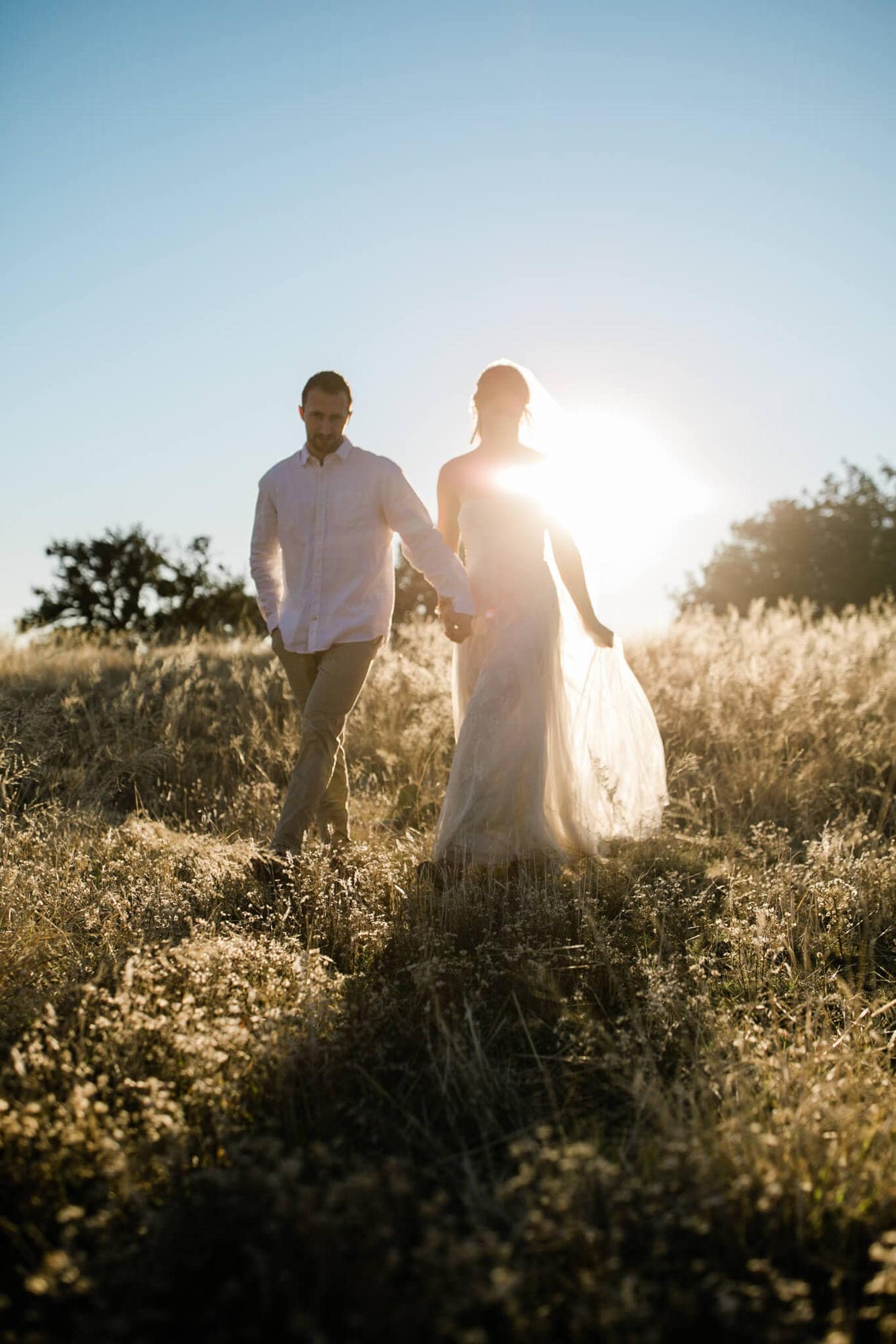 Silhouetted by the bright sun behind them, the bride and groom walk through tall dried grass.