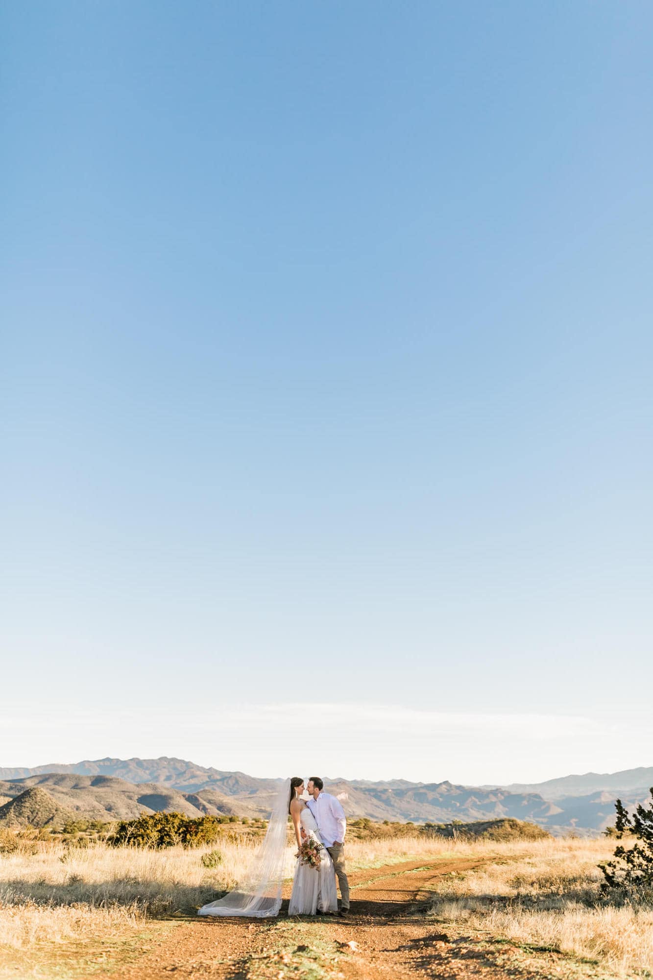 With the bright blue mountain sky behind them, the groom hugs the bride. 