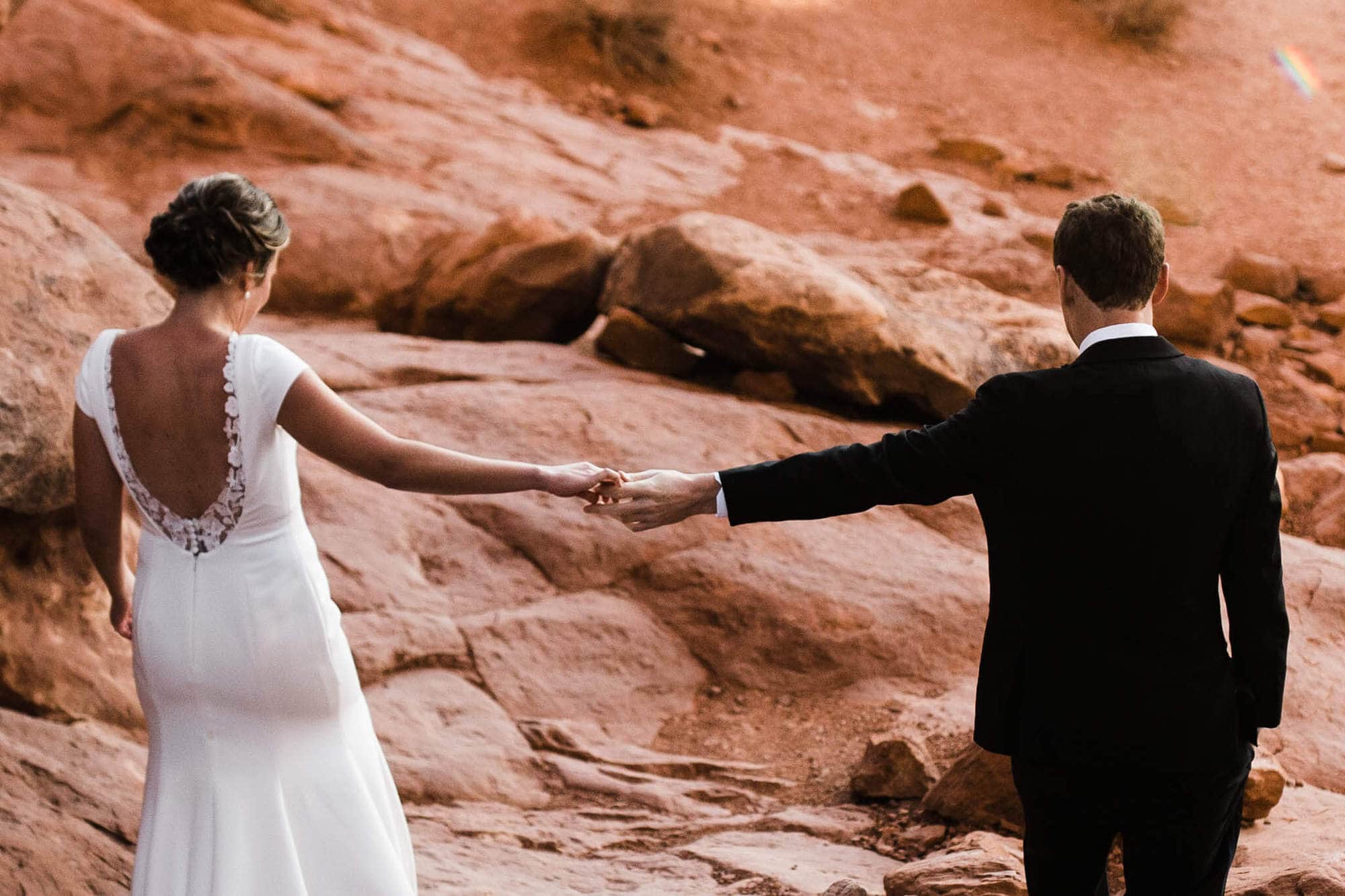 Bride and groom hold hands as they walk along the red rocks.