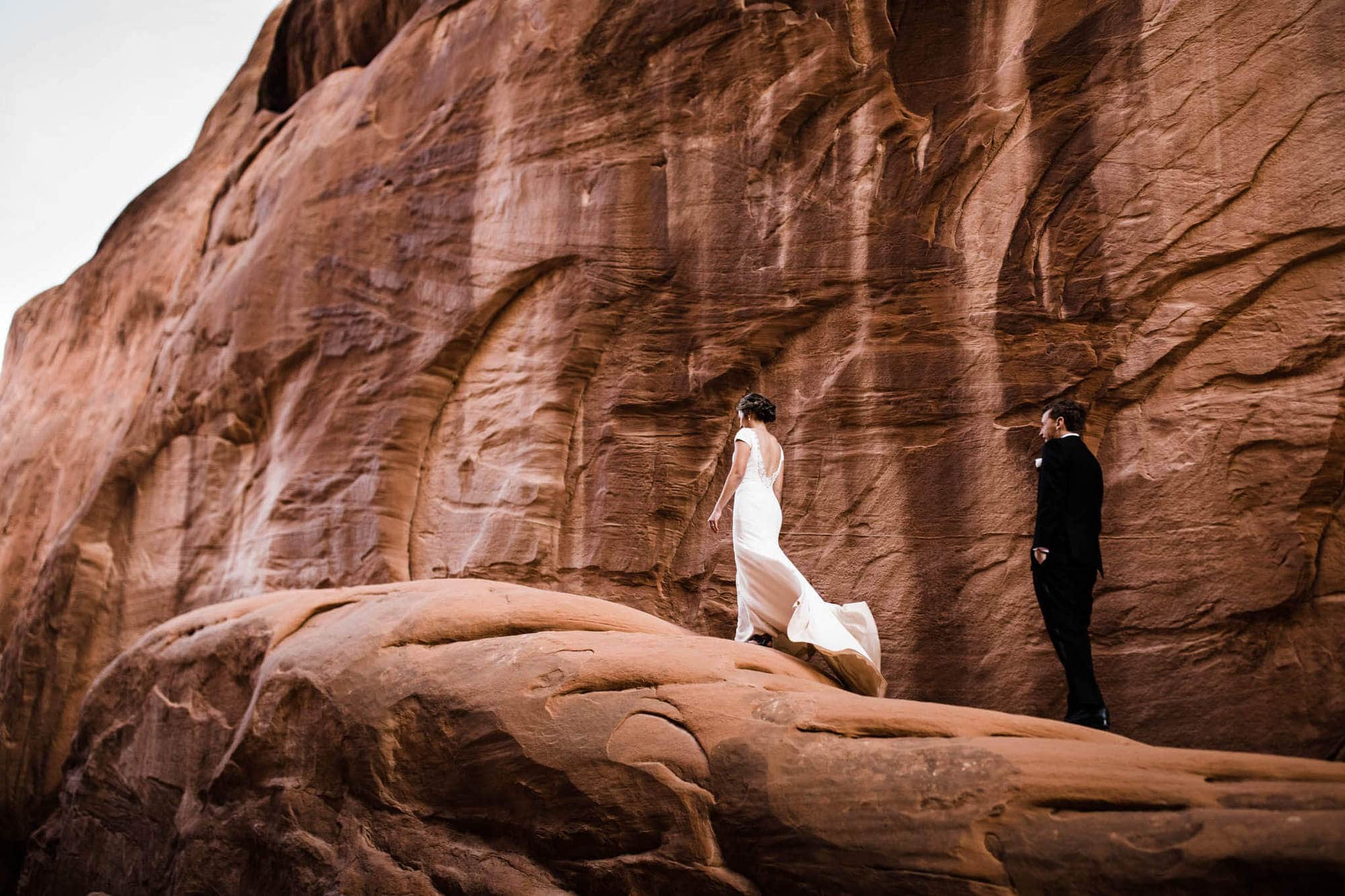 Couple stands on a ledge in arches national park as the wind catches brides dress.