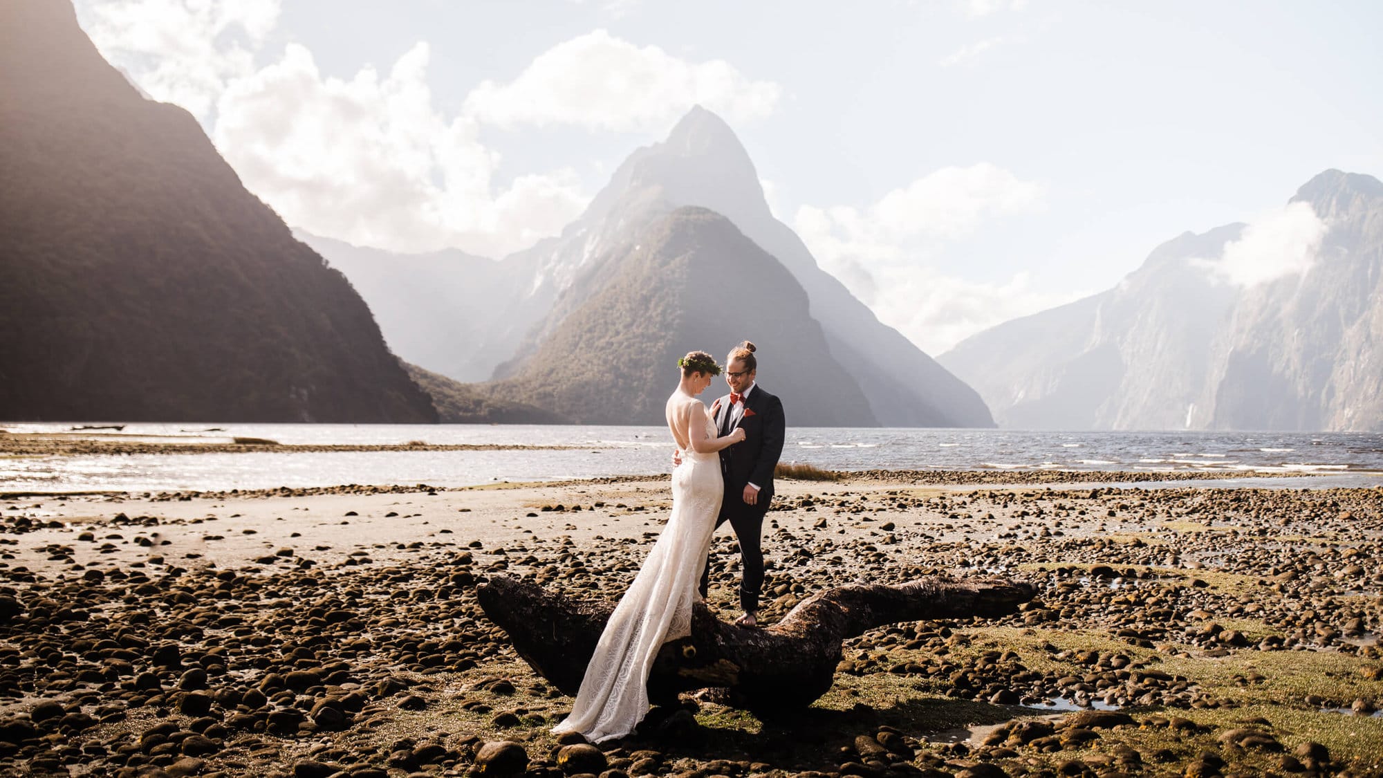 Couple stands on a log on the beach during their MILFORD SOUND Elopement. 