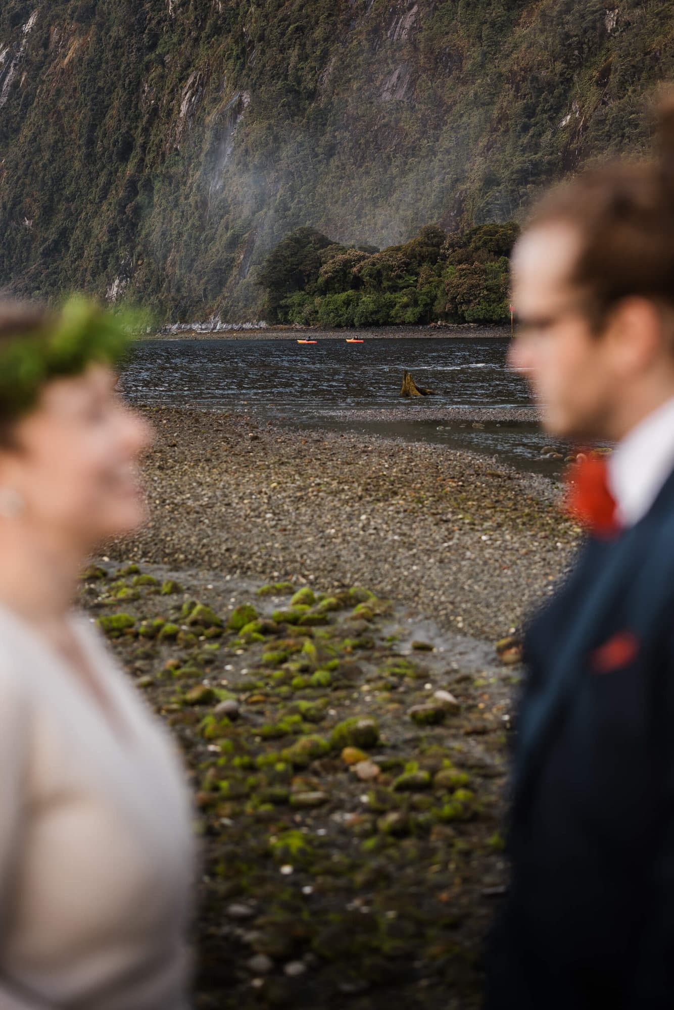 A couple looks at each other during their Milford Sound elopement while two kayakers paddle by. 