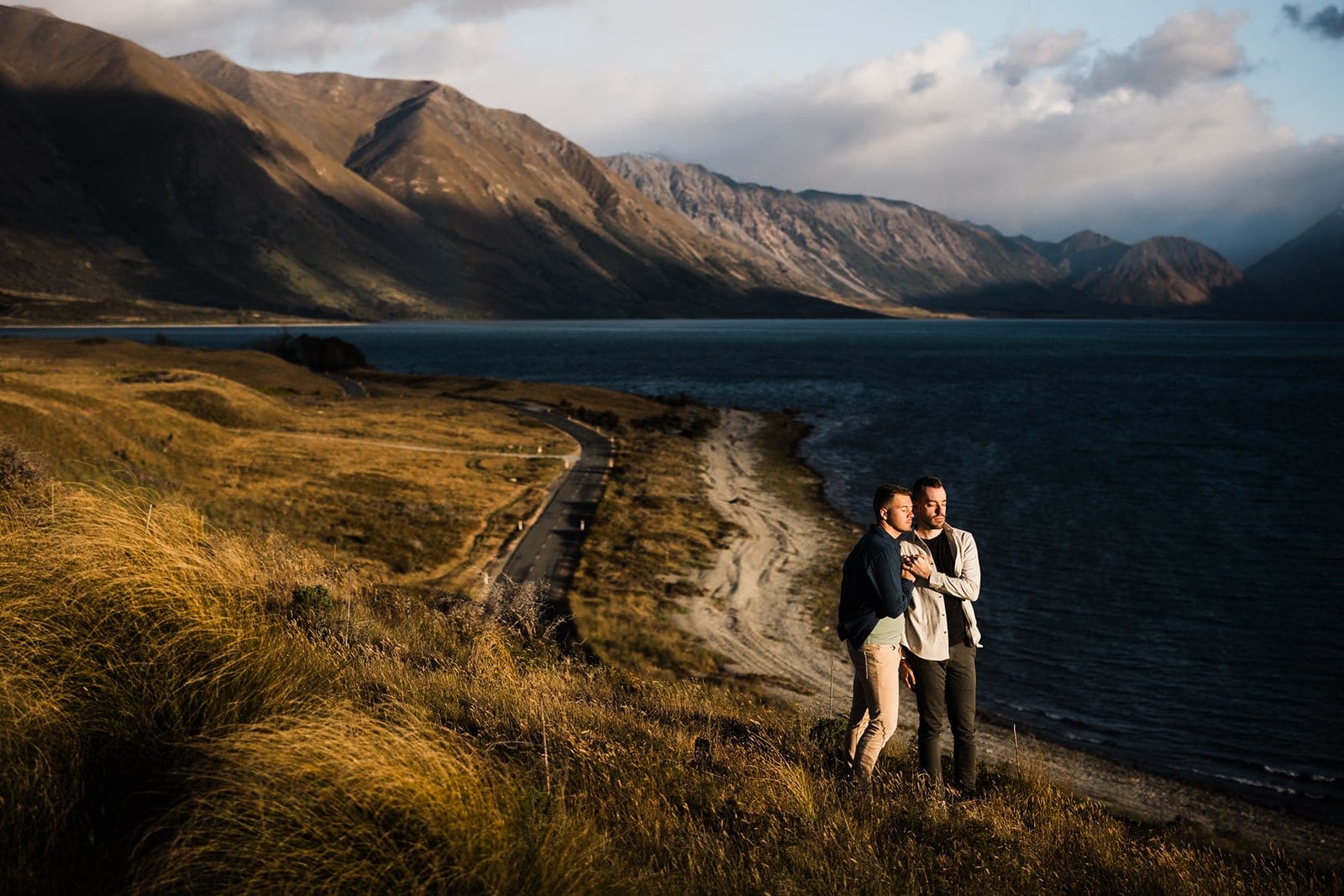 Two men hug while looking out over the water during their adventure photo session in New Zealand