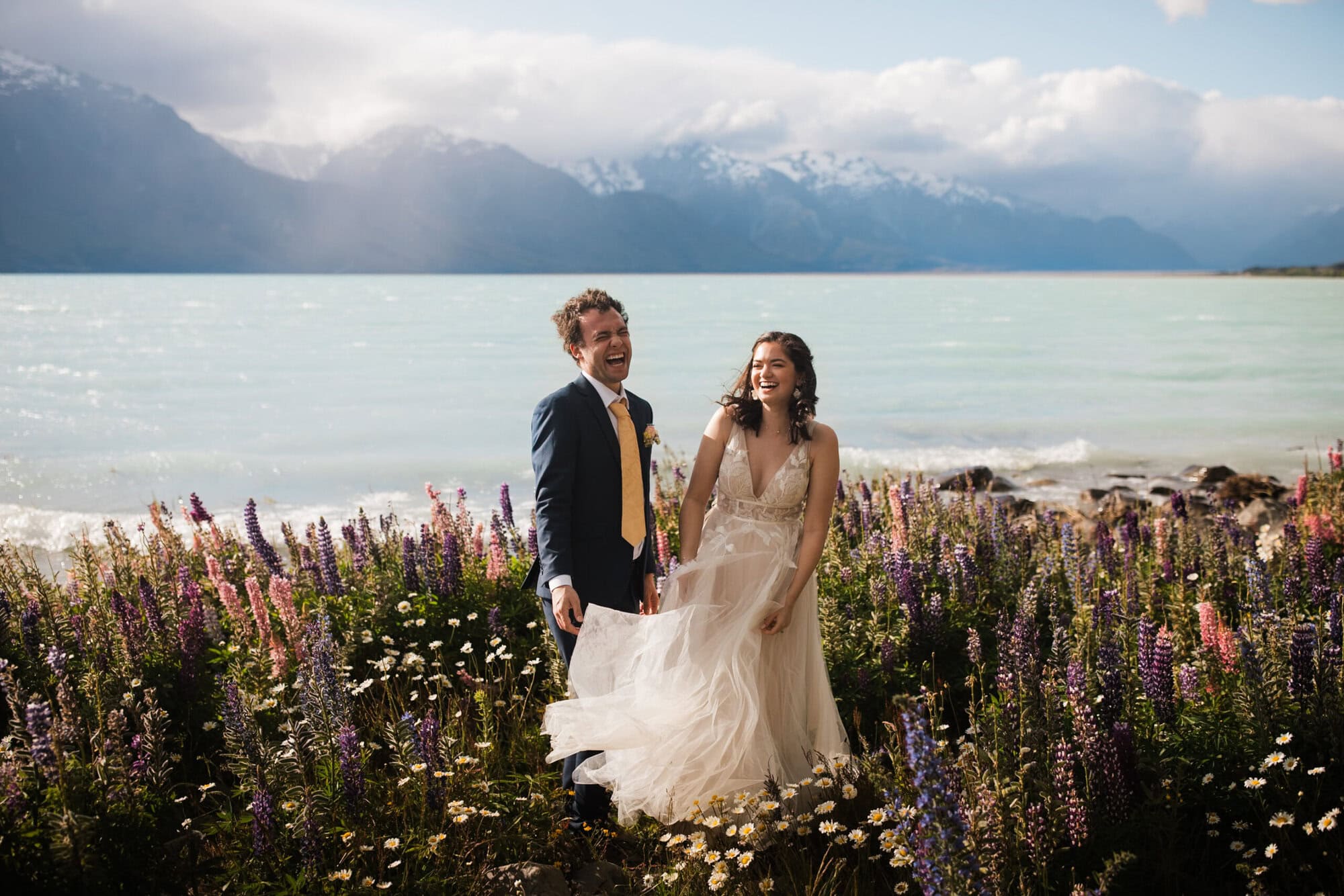 A bride and groom stand amongst a field of flowers with a lake and mountains as their backdrop, laughing as the brides dress catches in the wind. 