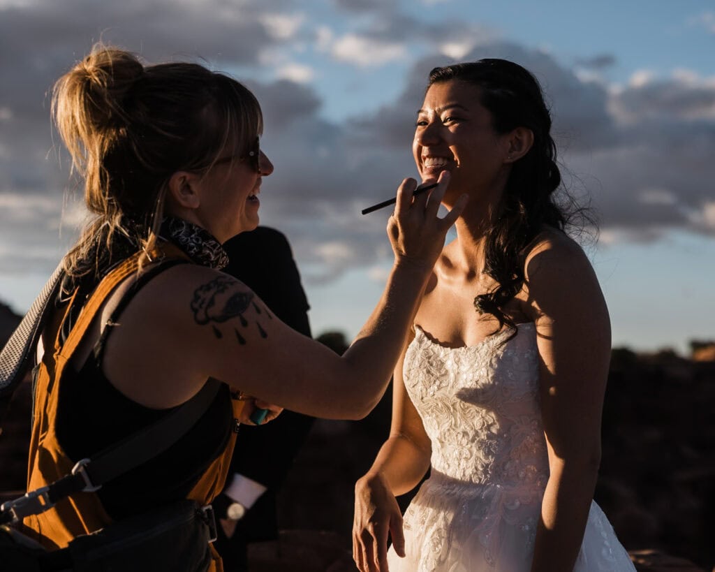 A smiling bride has her makeup adjusted by her makeup artist on her elopement day. 
