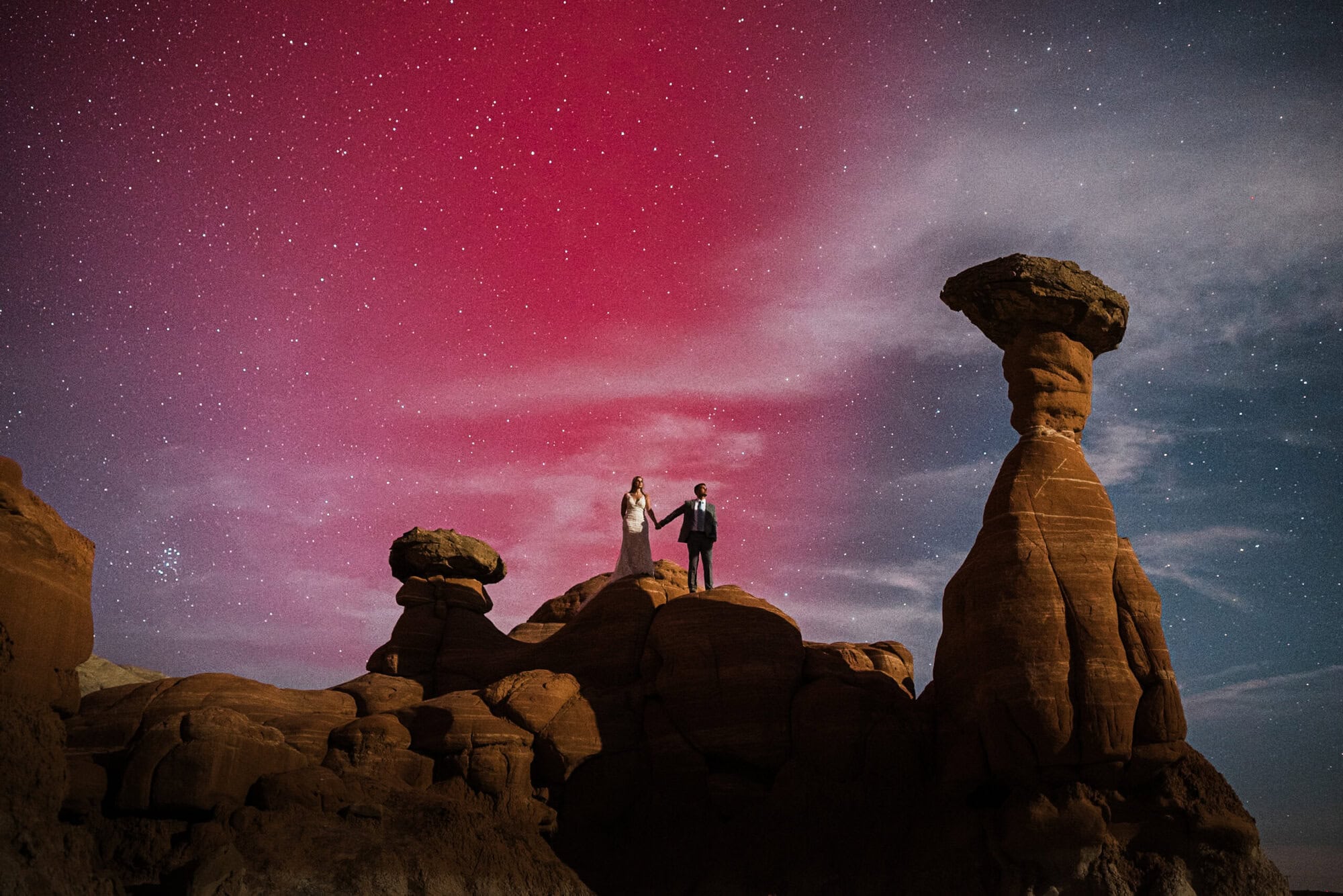 A bride and groom stand in the desert holding hands, the stars sky above them turned pink by a rare appearance of the Northern Lights. 