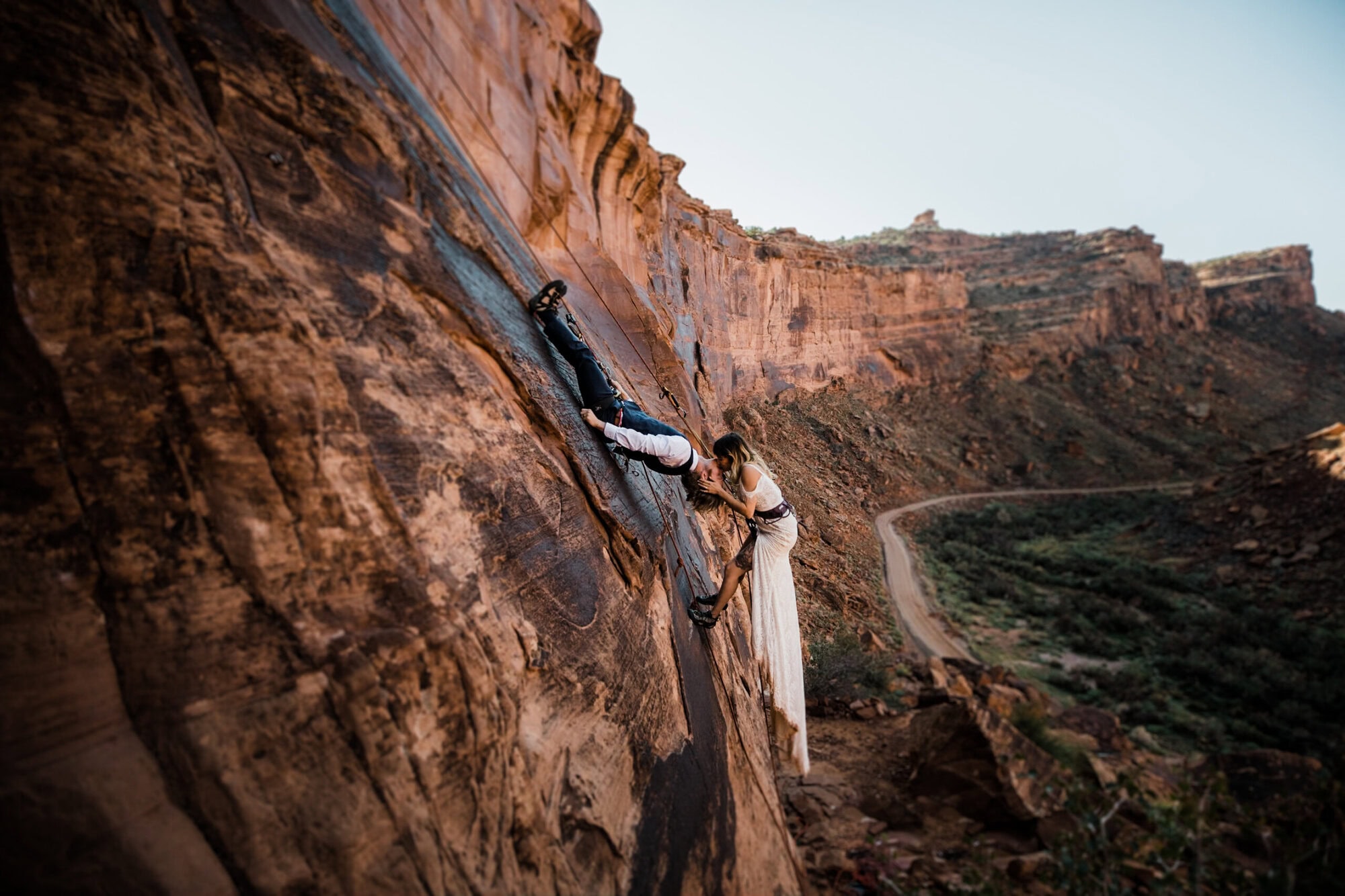 A bride and groom rock climb in Moab on their elopement day, the groom laying upside down and kissing his bride. 