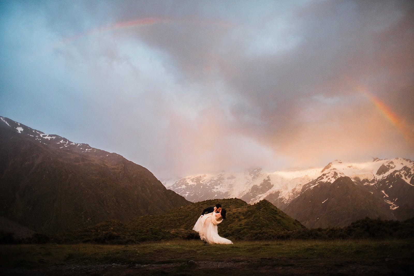 newlyweds kissing with a rainbow and mountains in the background in New Zealand, one of the best winter elopement destinations.