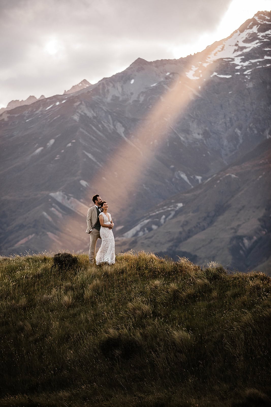 newlyweds standing on a grassy hillside with a spotlight shining on them and mountains in the background.