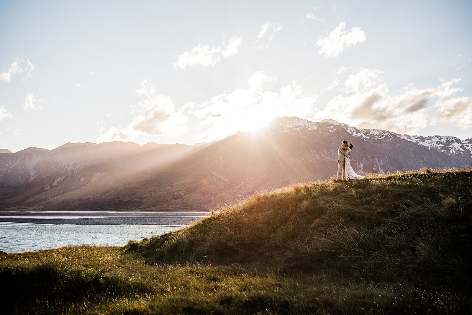 newlyweds kissing on a hillside with a rocky beach and mountains behind them in New Zealand, one of the best winter elopement destinations.