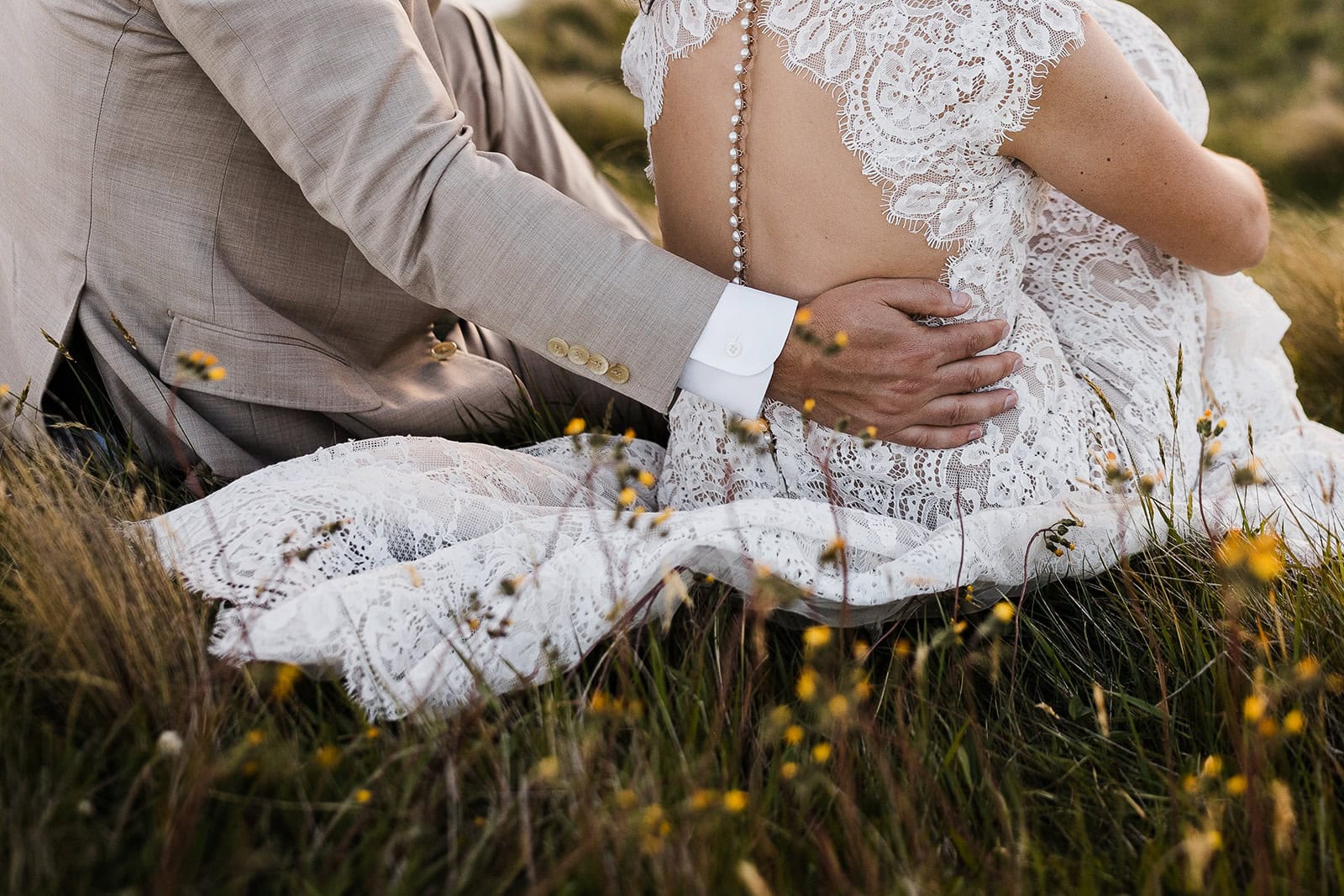 the groom holding the small of his wife's back while sitting in a field of wildflowers.