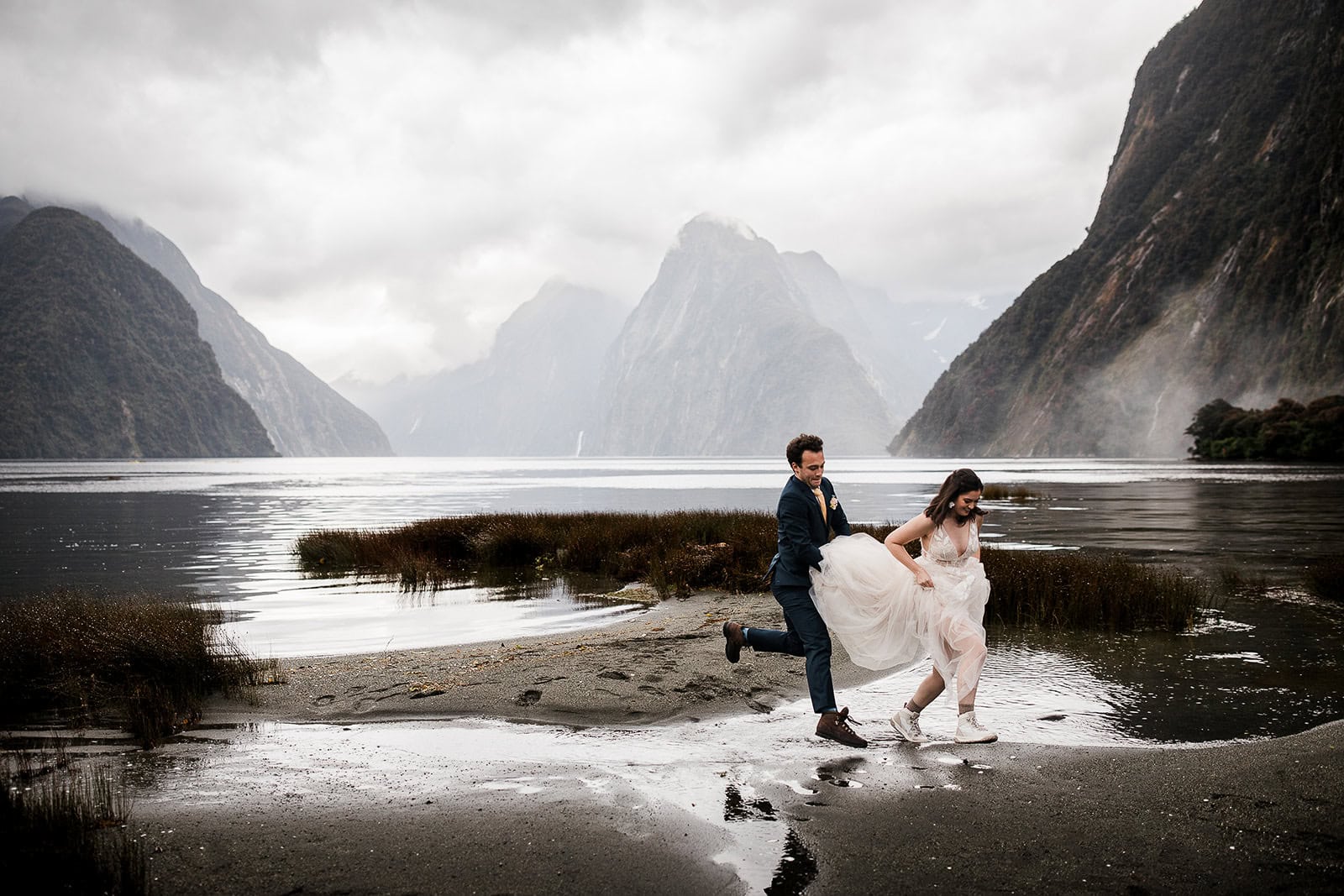 newlyweds frolicking on the beach at Milford Sound in New Zealand, one of the best winter elopement destinations.