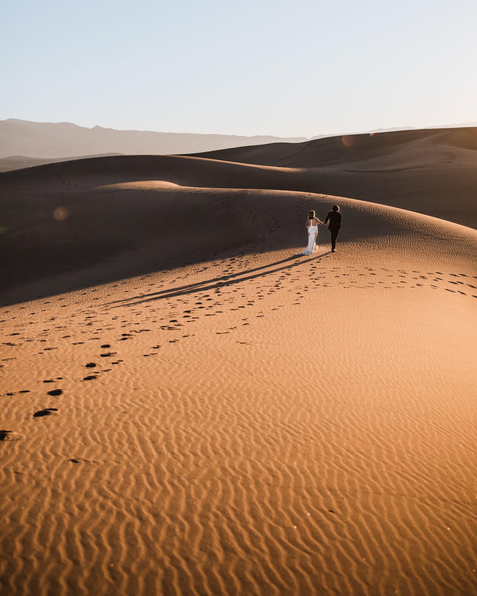 A couple on their elopement day walk hand in hand along the sand dunes towards the sunrise